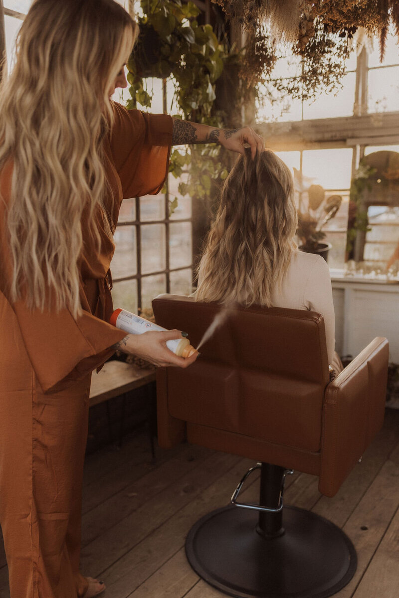 The hair extension specialist is working with her blonde client in Dallas, Texas. Behind them the salon has small windows and is decorated with green leaves on the ceiling.