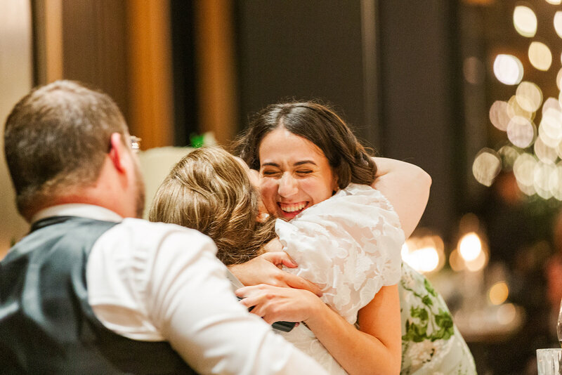 Wedding guest smiling with eyes closed hugging the jewish bride as she is seated at the sweetheart table with her scottish groom with his back to the camera