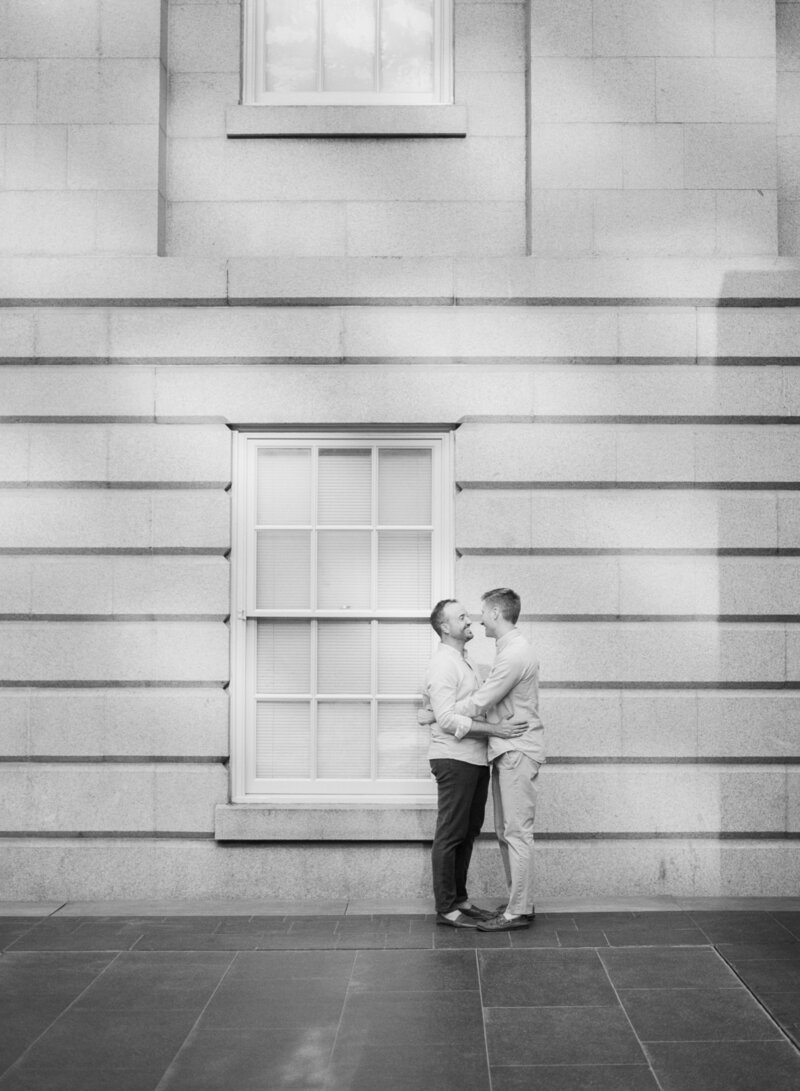 Bride and groom share a joyful moment; he kisses her cheek. She smiles brightly, holding a colorful bouquet. Autumn leaves create a warm, glowing backdrop.