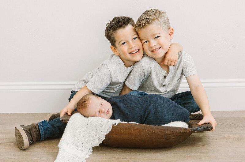 Two older boys look down at their newborn sibling who is sleeping in an antique wooden dough bowl.