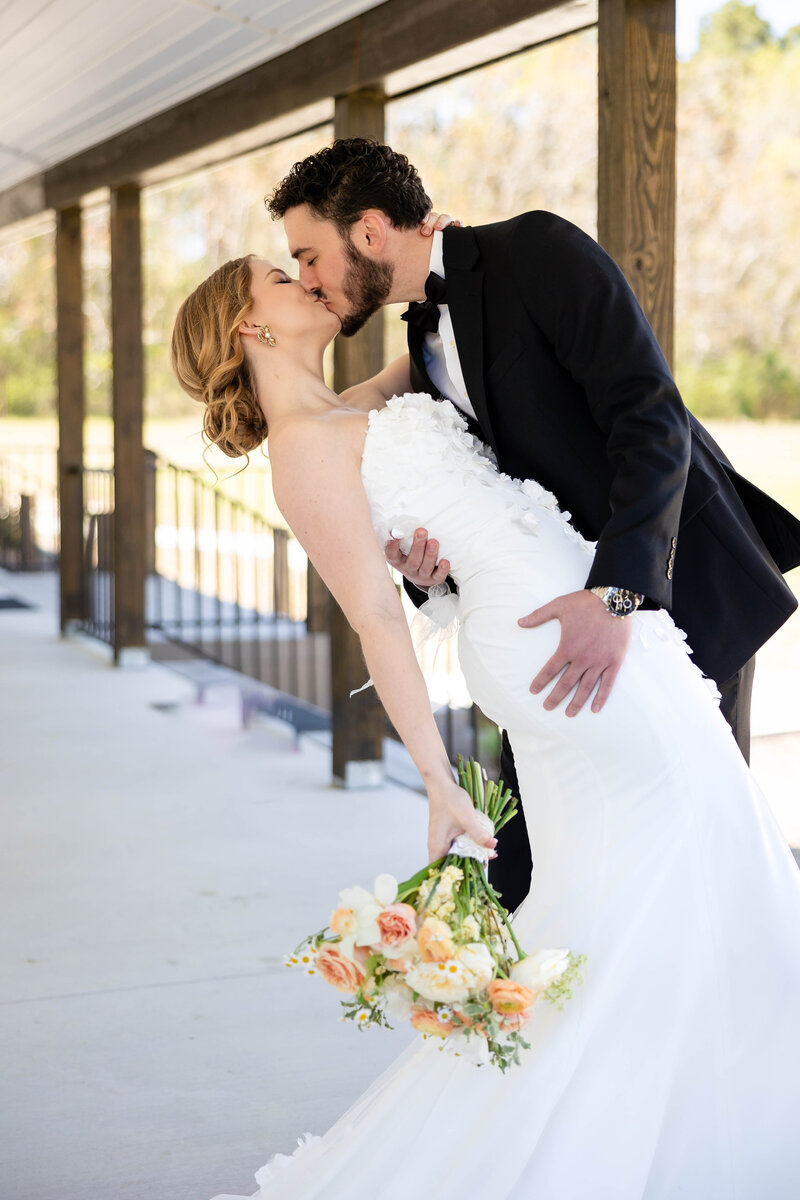 Bride and Groom on White porch with brown wooden columns.  Bride and Groom in a kiss while groom dips bride.  Bride dressed in a strapless fit and flare gown with white flowers attached to the dress.  Groom in a black tux with bowtie.