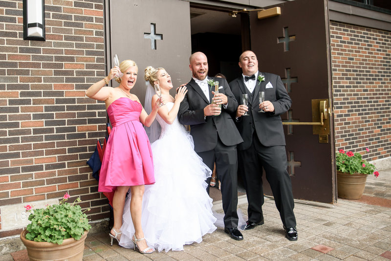 A couple and their friends celebrate their wedding by popping a bottle of champagne outside of the church