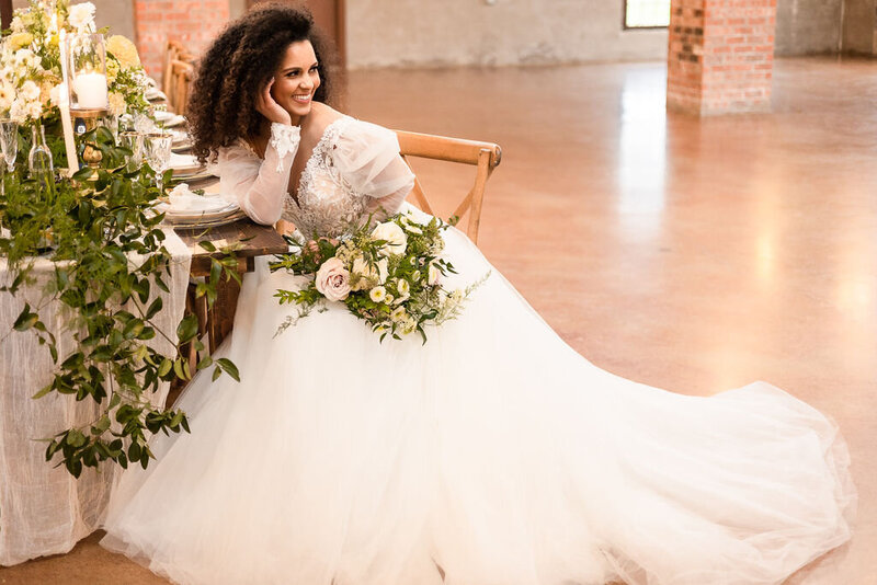 Bride in a tulle gown seated beside a candlelit reception table with lush greenery and blush florals in an industrial space.