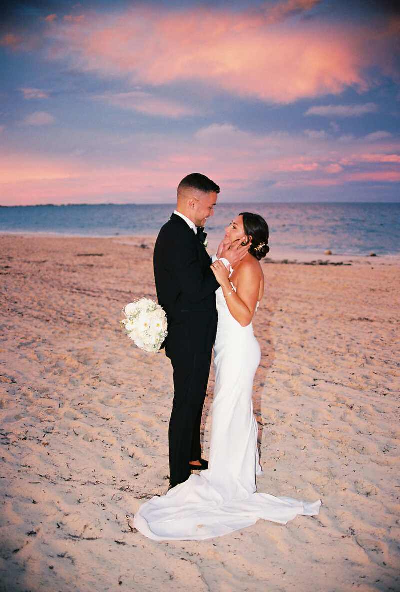 A bride and groom stand facing each other on a sandy beach at sunset, the bride holding a bouquet and the ocean behind them—captured beautifully by a Dallas wedding photographer.