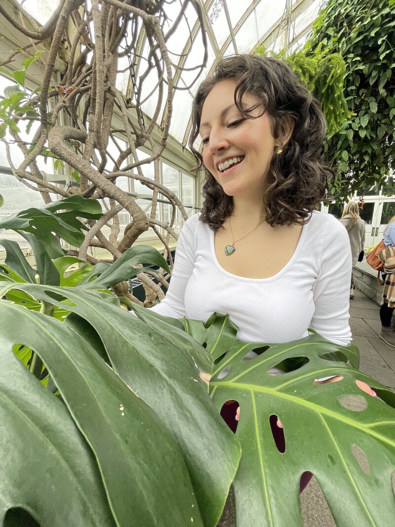 A wellness coach with brown, short, curly hair stands in a lush garden, looking down and smiling at a huge monstera leaf. The natural surroundings and the gentle smile on her face create a sense of calm and tranquility. The image evokes feelings of relaxation, self-care, and mindfulness, making it perfect for anyone interested in wellness and personal growth.  [Nicole QW]