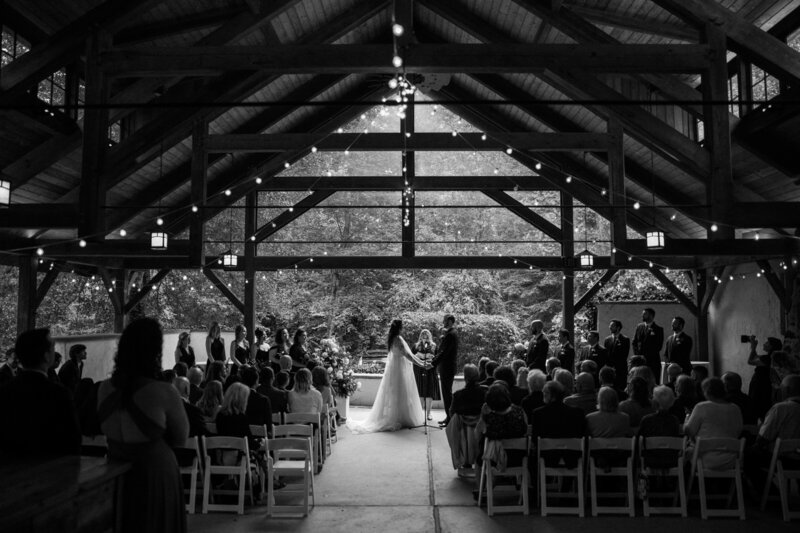 A black-and-white wedding ceremony inside a rustic venue, with guests seated and a lush outdoor backdrop visible through large windows.
