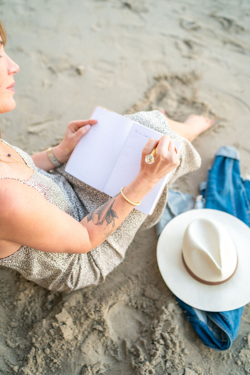 A woman life coach sitting on the beach writing in her journal to share her story.