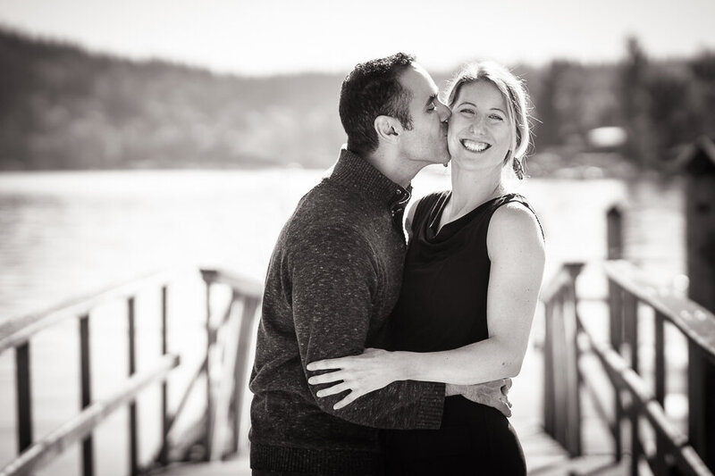 Parents kissing during a holiday family photoshoot session with their three children. Photograph shows a beautiful glowy, misty backdrop along a private path in Redmond WA.