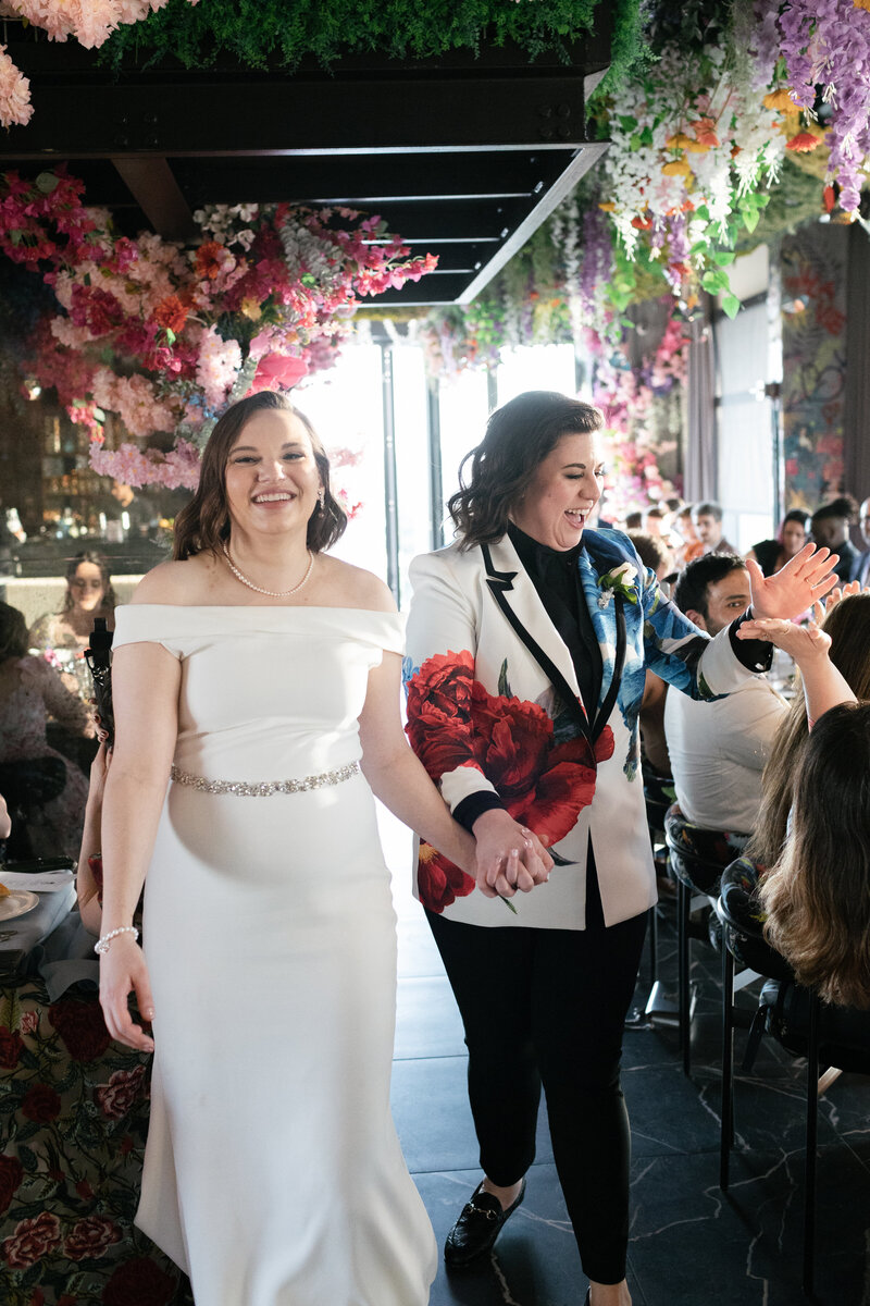 lesbian brides walking into their wedding reception smiling at the camera and high-fiving a guest walking by
