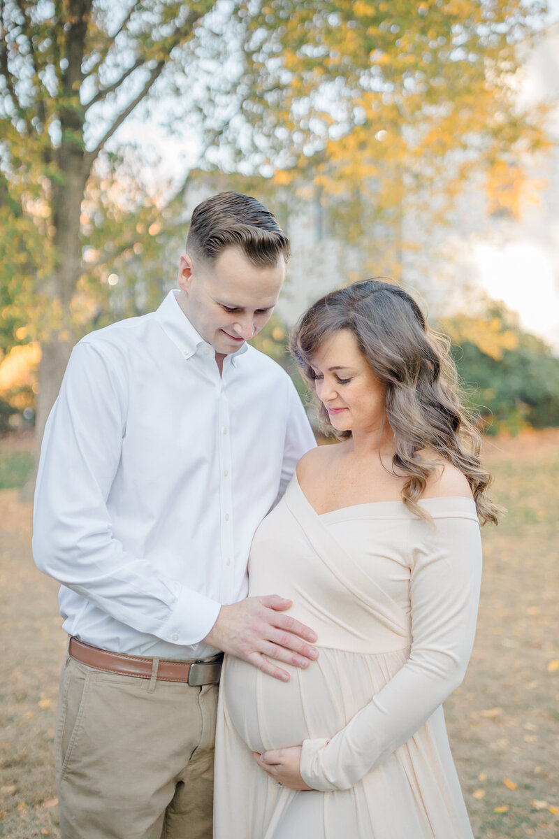 Amidst the golden hues of autumn, a husband and wife stand together, gazing lovingly at her growing belly, filled with anticipation and joy for the new life they will soon welcome