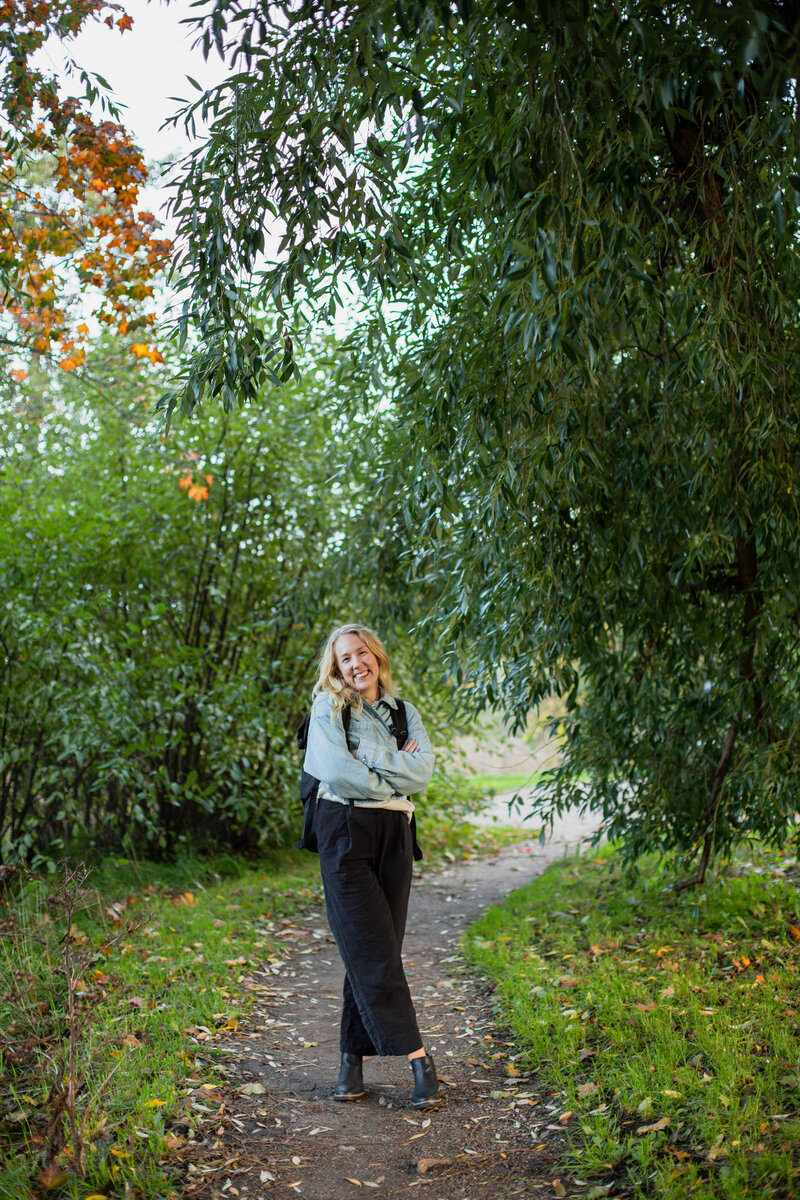 mielen paremminvoinnin ja kehomieliyhteyden valmentaja  seisoo metsäpolulla puiden keskellä ja hymyilee