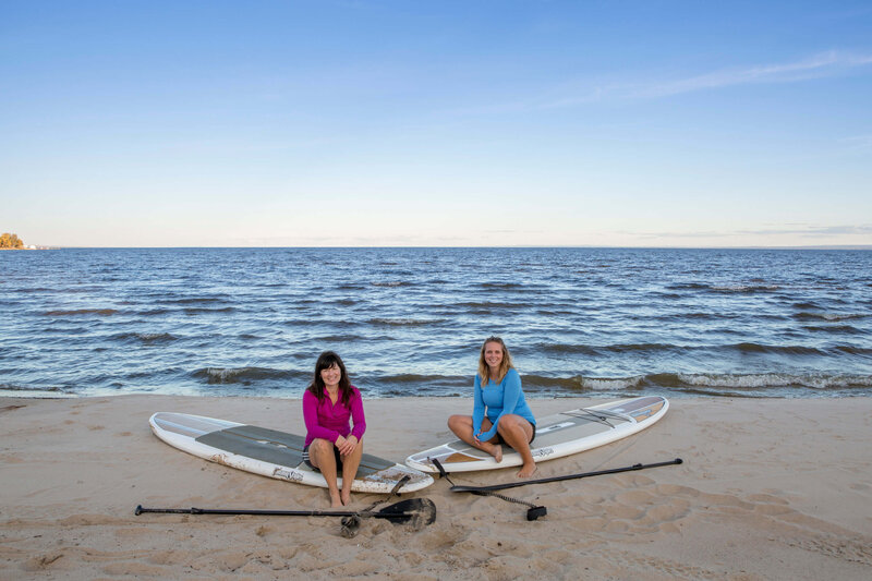 2 women sit on their paddle boards on the  beach