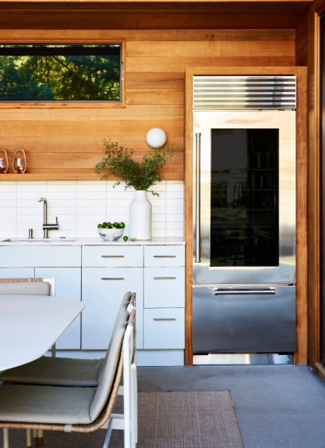 Hingham interior designers create an outdoor kitchen for a new construction home in Hingham, Massachusetts, featuring light grey metal cabinets, white counters, and red cedar walls and ceilings.