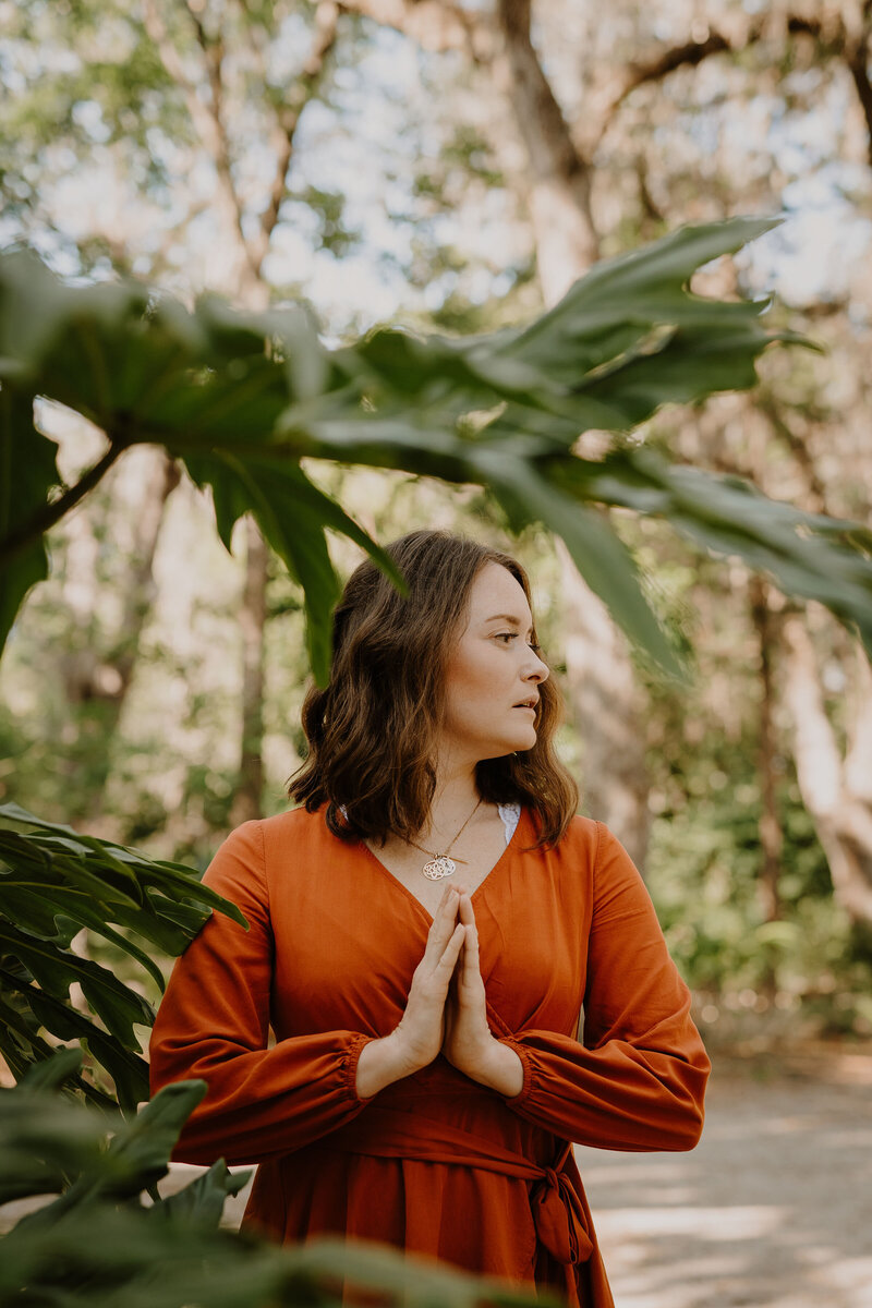 Woman in orange dress stands in prayer among leafy green plants.