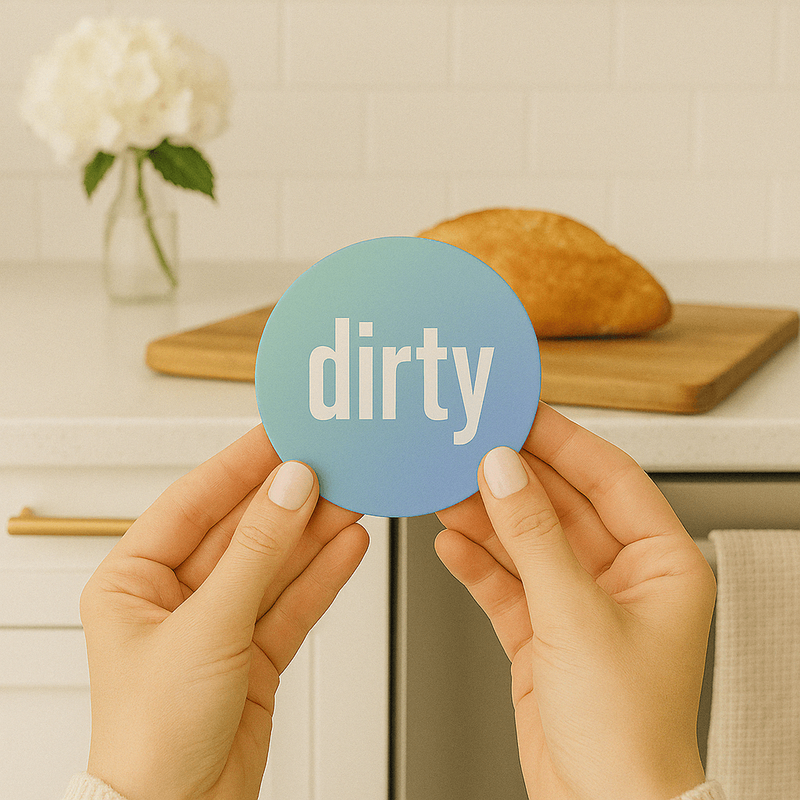 Two hands hold a blue, round magnet with text that says "dirty" in lowercase, bold, white letters with a elegant, minimalistic kitchen in the background