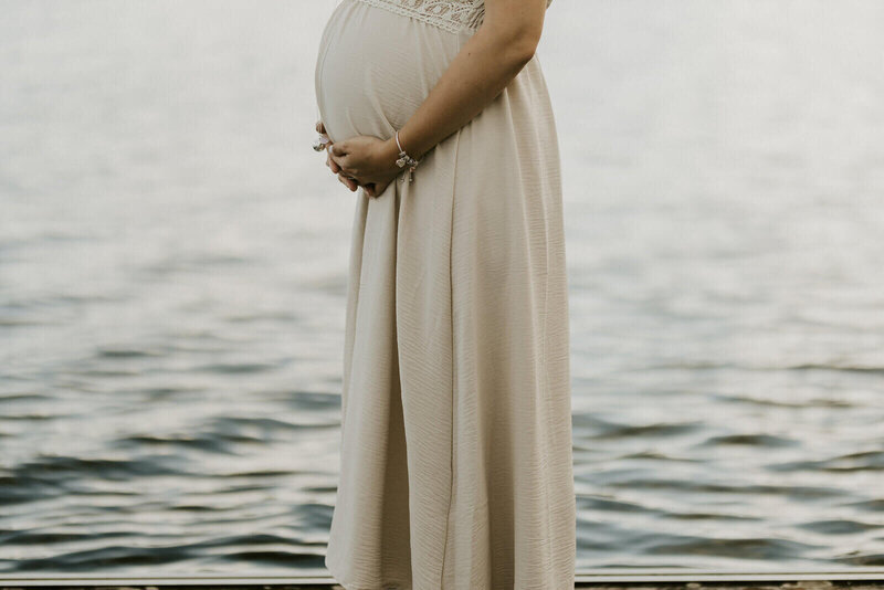 Ventre arrondi d'une femme enceinte de profil, en robe longue blanche, au bord de l'eau, capturée par Laura, photographe grossesse en vendée.