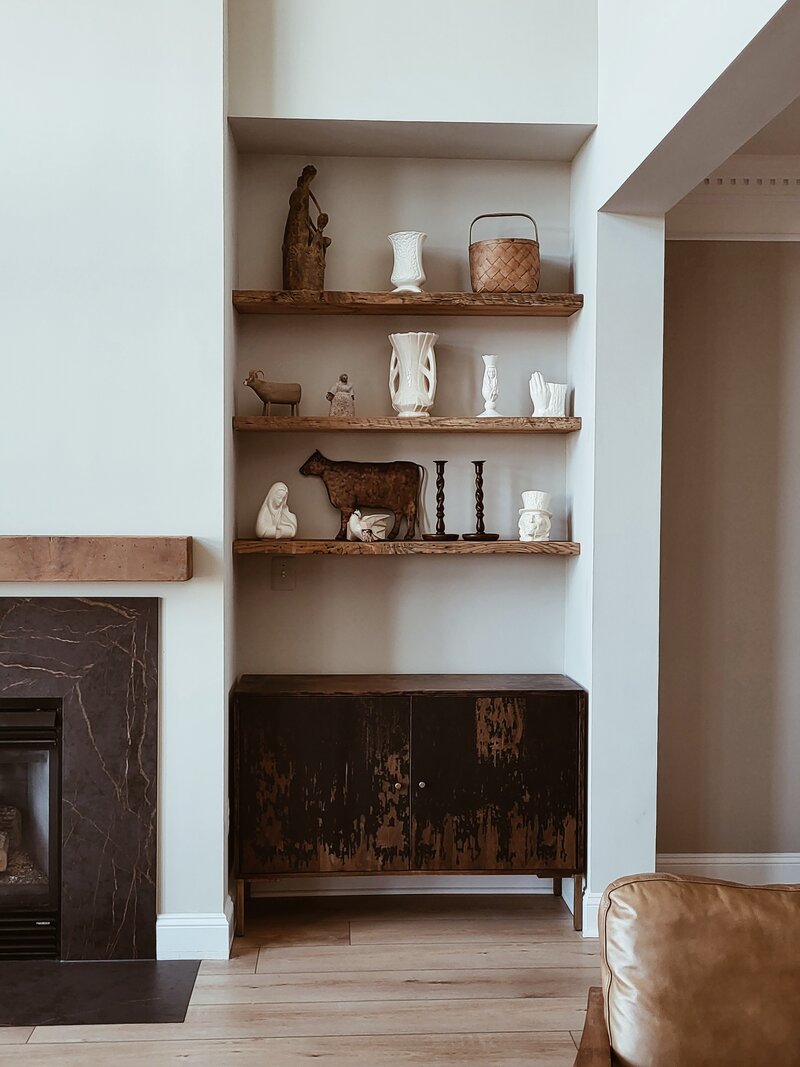 side shot of a fireplace with black surround face front to the fireplace with a wooden mantle a nook beside it with antique raw wood shelves and an artistically painted cabinet that matches the wood tones of the shelves and the fireplace surround