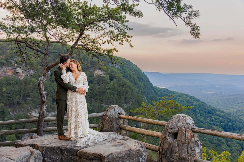 A bride and groom stand on a rock overlooking a mountain view while the sun sets. The bride is standing in front of the groom and is leaning her head back on his shoulder while gently holding his face and looking at him. The groom has his arms wrapped around the bride and is looking down at her.
