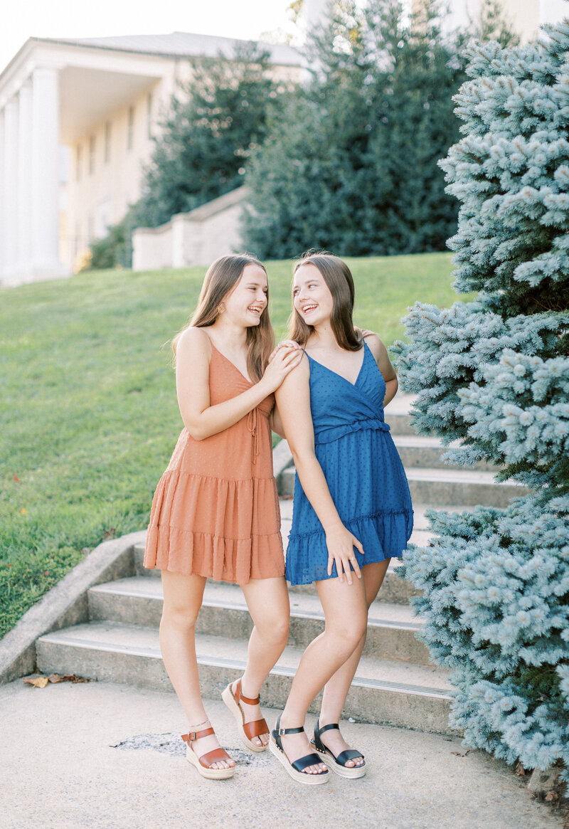 Twin girls stand near a evergreen bush and concrete steps and laugh at each other playfully.