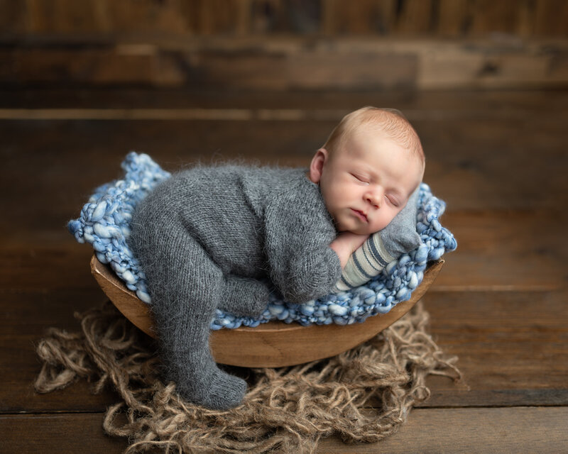 Peacefully posed in a wooden bowl with soft blue knits, this newborn boy is dressed in a fuzzy grey sleeper – a cozy rustic setup in our Paris, Ontario studio.