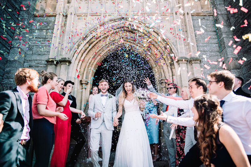 A couple on their wedding day walking out of a church with confetti being thrown over them captured by Devon Wedding Photographer