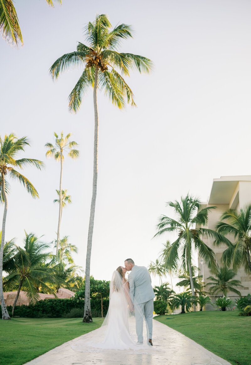 A bride and groom kiss on a pathway with palm trees at their tropical destination wedding