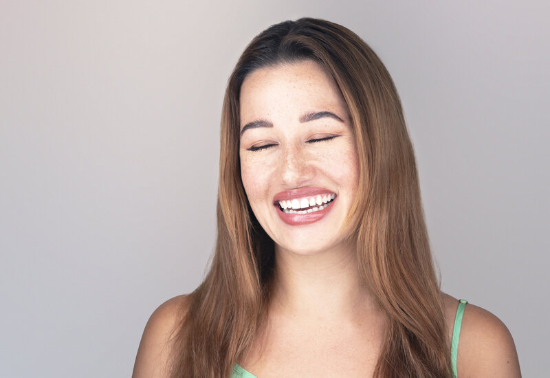 Woman in green tank top with long brown hair poses for a studio headshot. Her eyes are closed and she is laughing. Philadelphia and Main Line Headshot Photographer.