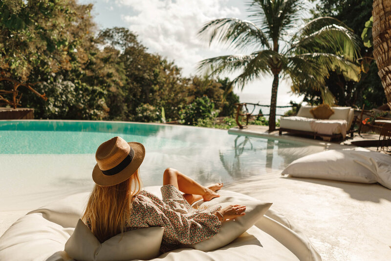 Woman wearing a straw hat and floral dress relaxing on a lounge chair by a tropical pool surrounded by lush greenery and palm trees.