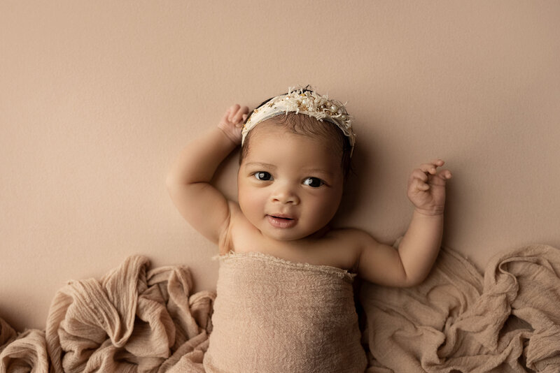 Newborn photoshoot in top London, Ontario studio. Baby is laying on her back with big brown eyes looking at the camera. Her belly is draped in a tan fabric and she is wearing a coordinating delicate floral headband. Baby's arms are resting above her head.