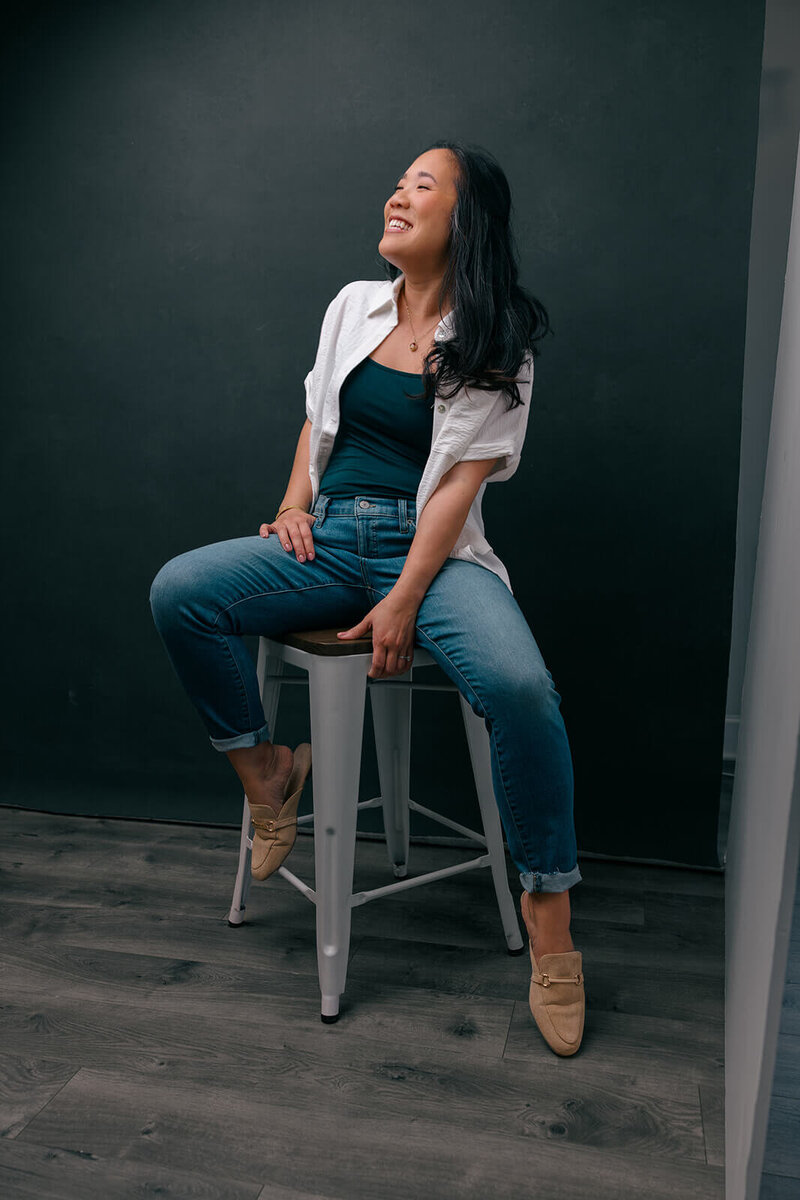 asian woman sitting on a stool, laughing freely who is wearing a green tank top with a white blouse and jeans