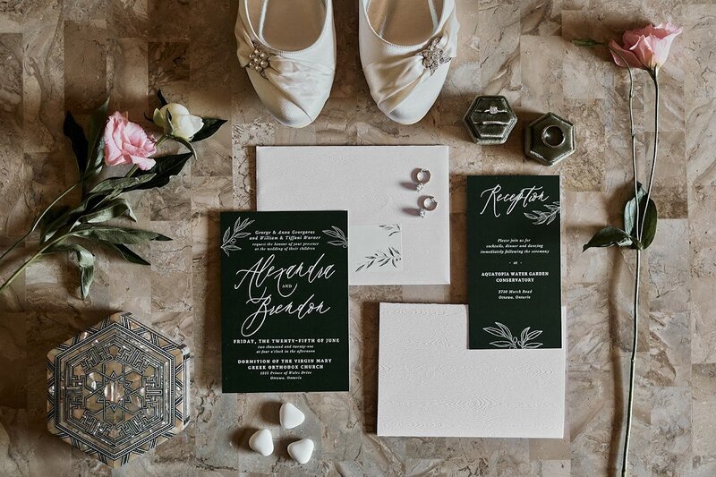 A modern minimalist forest green wedding invitation suite with olive branch line art sits on a table with florals, rings, the bride's shoes and an ornate ring box at a wedding at Aquatopia in Almonte, designed by Frid Events.