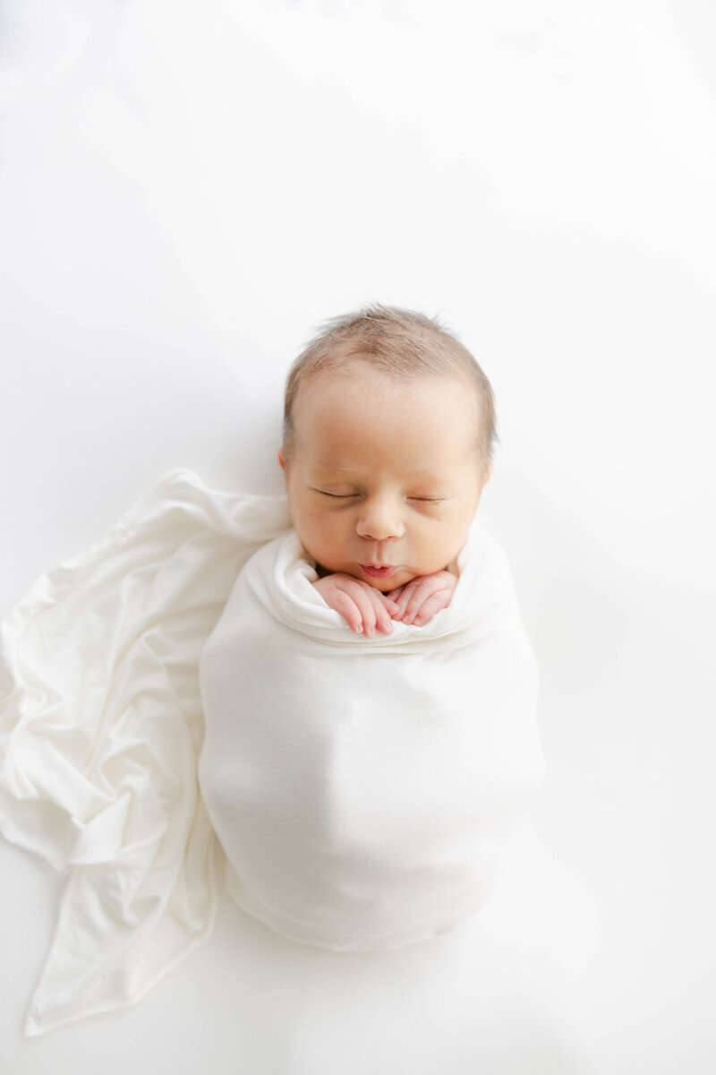swaddled newborn sleeps with his lips pursed and his hands holding the white swaddle Indianapolis portrait photographer session