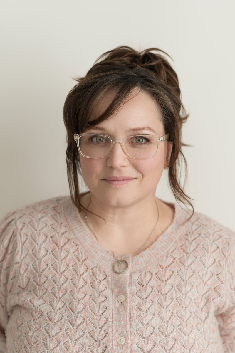 a woman smiling at the camera, she is wearing a pink sweater and wearing clear plastic glasses and has brown hair