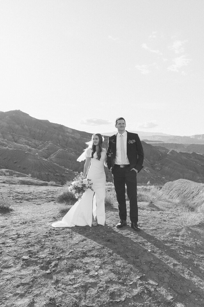 A black and white photo of a bride and groom. The bride is holding the groom's arm and they are looking in opposite directions, not at the camera. Behind them is the stunning landscape of Capitol Reef National Park located in Torrey, Utah.