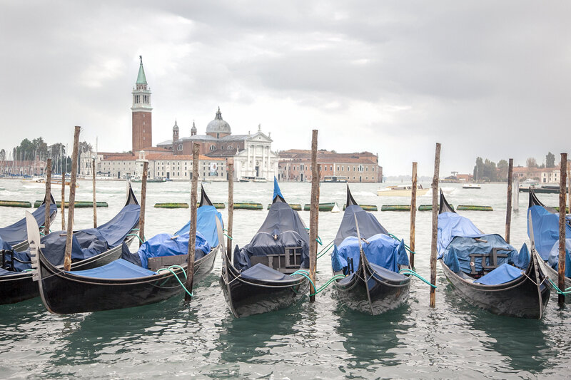 A row of gondolas with blue covers floats on a canal in Venice, Italy. In the background, the San Giorgio Maggiore island is visible, featuring historic buildings and a prominent bell tower under a cloudy sky.