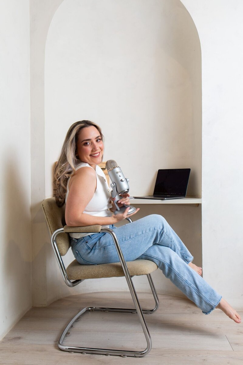 Merlyn Asencio sitting at her desk with a mic in hand and a warm smile, exuding approachability and professionalism as she engages with clients through her trauma therapy practice in Miami.