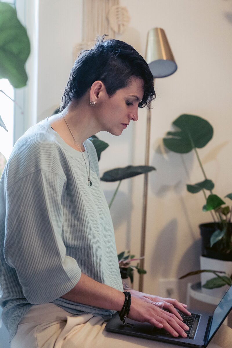 This image frames a white woman sitting and working on a laptop on their lap. The image is a side view angle with some house plants in the background