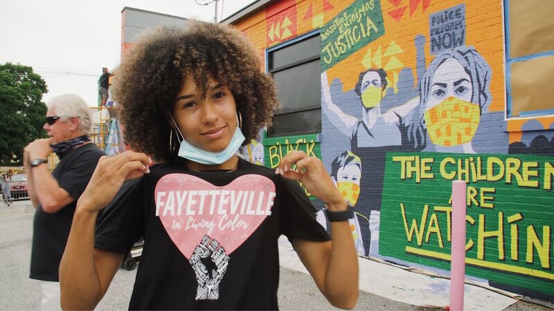 Woman wearing "Fayetteville in Living Color" t-shirt standing in font on black justice mural
