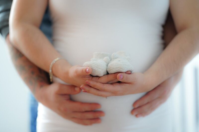 A pregnant woman tenderly holds small, knit baby boots in front of her belly, while her husband lovingly embraces her from behind. This heartwarming image captures the intimacy and joy of expecting parents, embodying the essence of our Portrait, Newborn & Maternity Photography service, where we celebrate life's most precious moments with warmth and artistry.