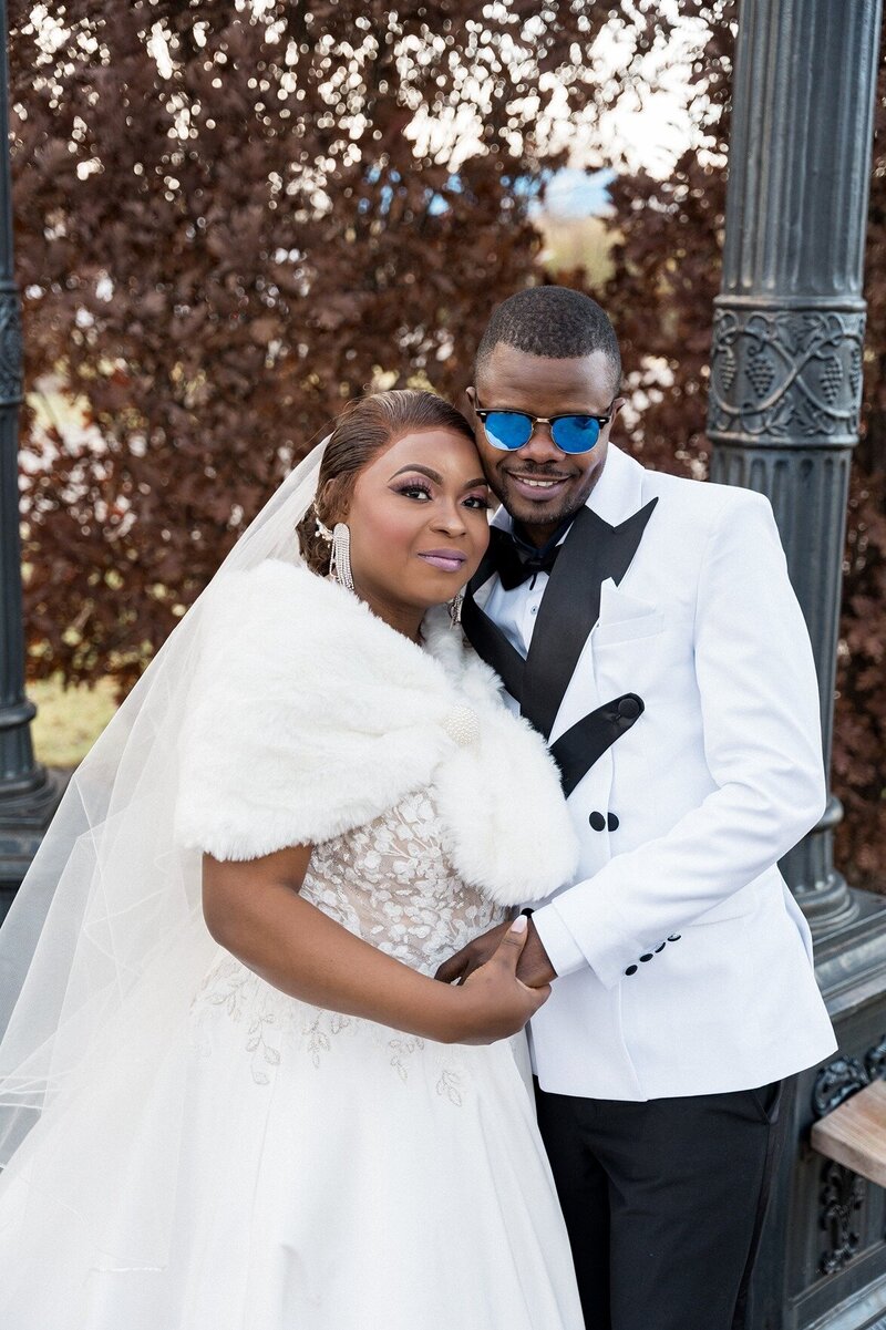 A bride and groom pose closely together in an outdoor setting. The bride, adorned with a glamorous makeup look and wearing a white fur shawl over her lace wedding dress, smiles beside the groom, who sports stylish blue sunglasses and a sharp white tuxedo with black lapels. They stand in front of a blurred backdrop of autumnal brown leaves, adding a warm tone to the scene.