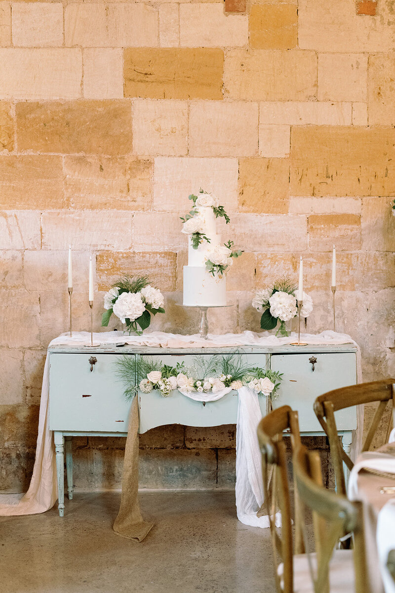 Elegant white tiered wedding cake with green accents on vintage sideboard, ideal for luxury Puglia celebration.