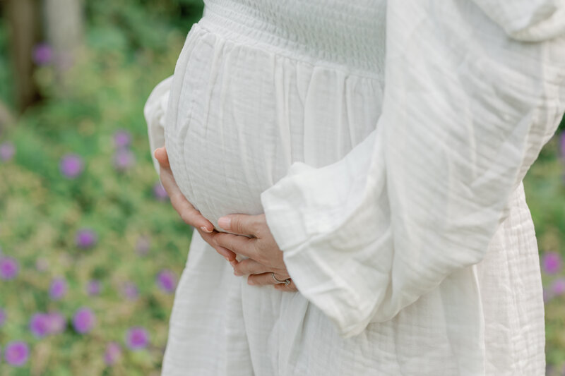A soft, dreamy maternity portrait featuring a mother-to-be cradling her baby bump in a flowing white dress. Surrounded by vibrant blooms at Alexander Muir Gardens in Toronto, this image captures the beauty and anticipation of pregnancy in a peaceful, nature-filled setting. Photographed by KCP Families.