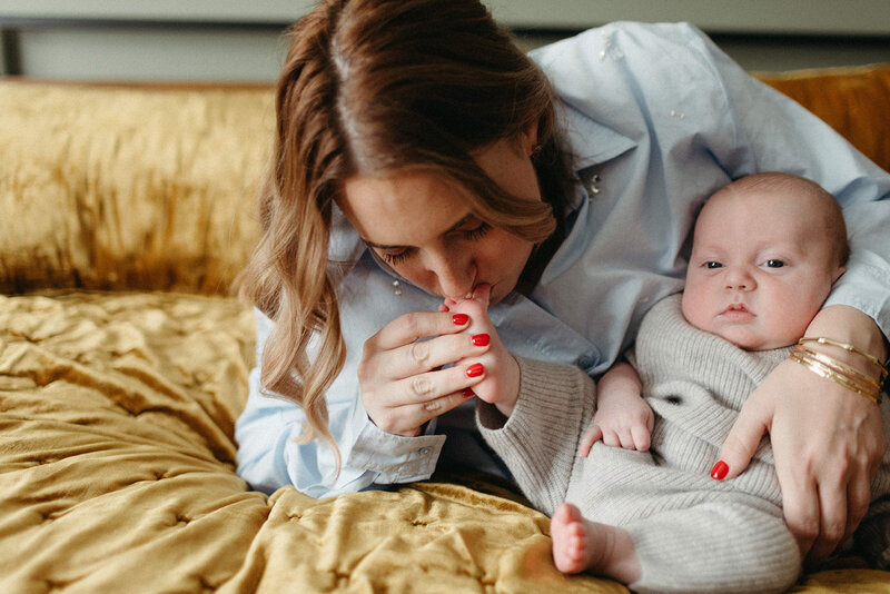 A woman lies on a mustard yellow blanket, gently kissing her baby's hand. The baby, in a gray outfit, is cradled in her arm. This tender moment, perfectly captured by a Toronto newborn photographer, exudes warmth and love.