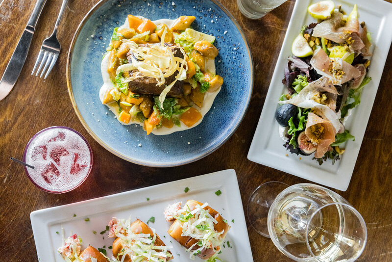 Elegant restaurant spread featuring a plated short rib dish with gnocchi and butternut squash, a prosciutto and fig salad, and lobster rolls on toasted buns. A glass of white wine and a sparkling cocktail accompany the meal on a rustic wooden table. Small business branding photography in Columbia, SC, showcasing fine dining, artisanal cuisine, and local restaurant branding.