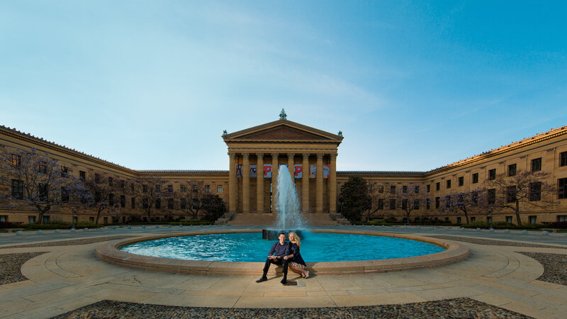 Engaged couple sitting at a fountain in front of the Philadelphia Museum of Art, sharing a candid and romantic moment.