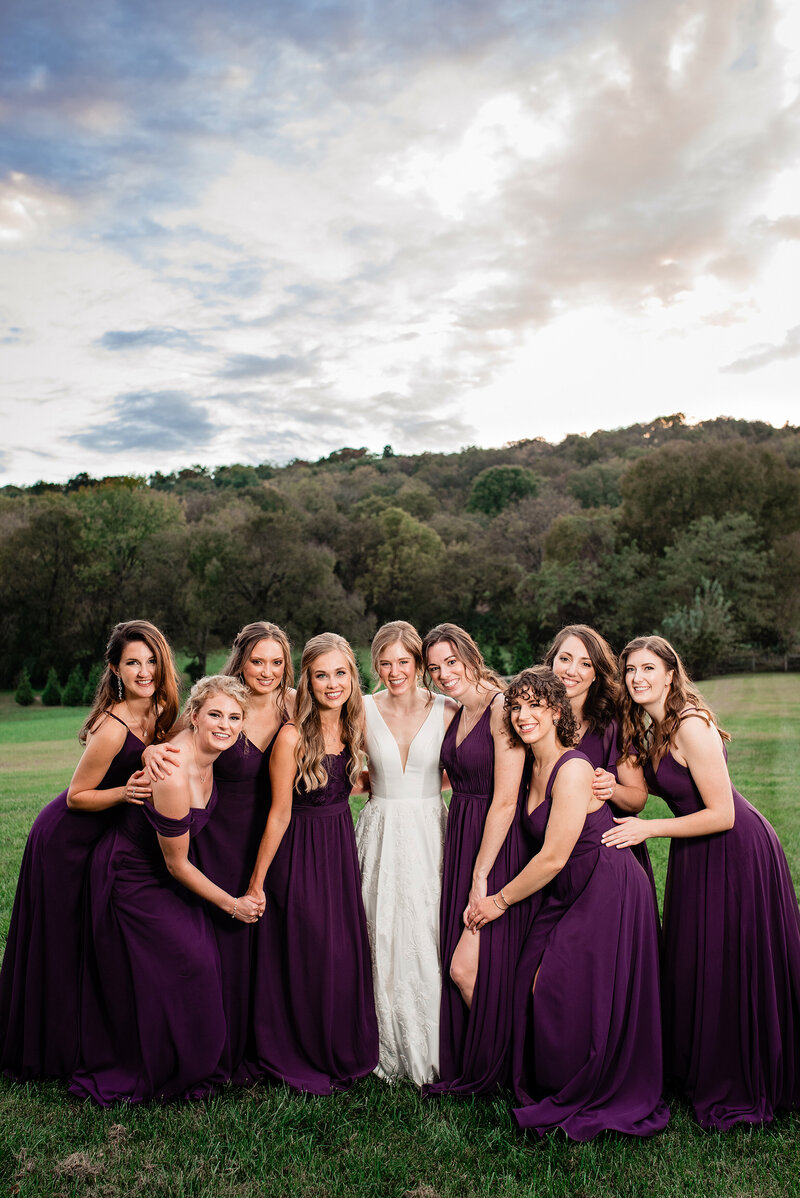 Bride surrounded by her bridesmaids all smiling at the camera, the girls are wearing purple dresses and they are outside in a grassy area with trees in the background at sunset