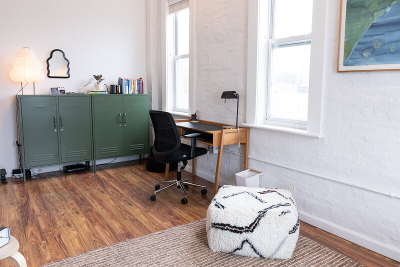 Wooden desk with black rolling chair facing two windows, green office cabinet with books atop and white ottoman with a black abstract pattern in an office with wood floors in Greenpoint, Brooklyn.