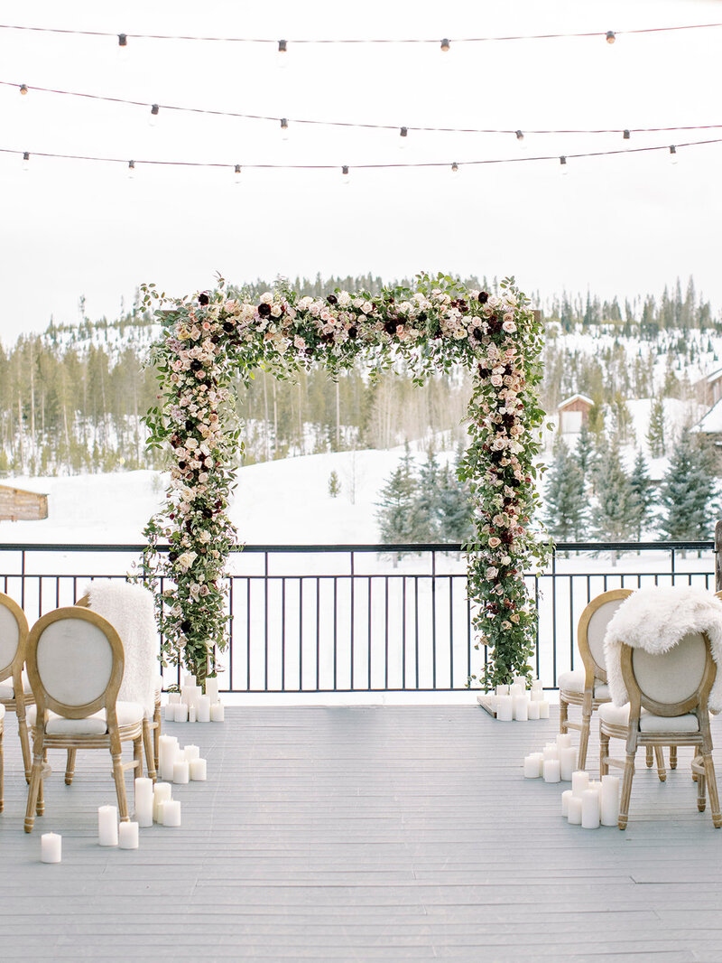 Empty snowy outdoor wedding aisle with white runner, gold chairs, and flower-covered archway.