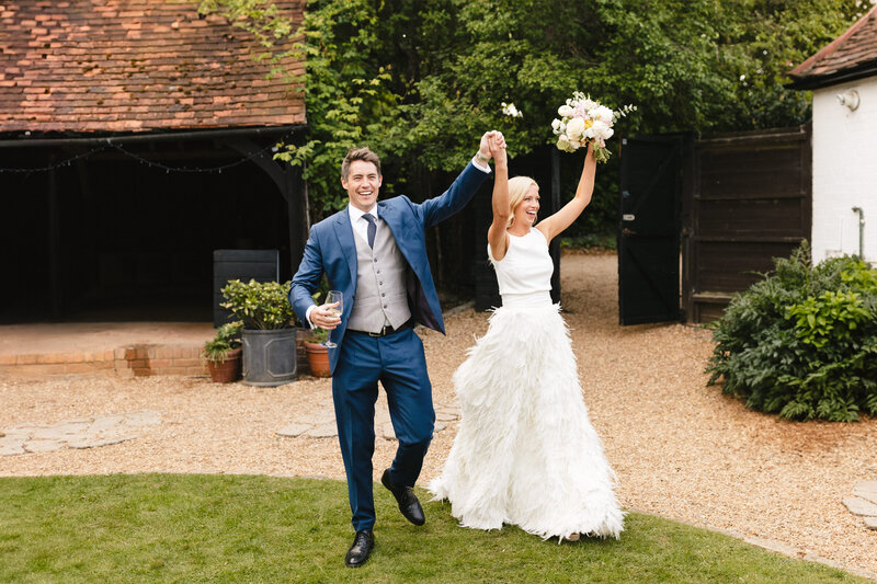 A newlywed couple joyfully walks outdoors on a lawn. The groom, in a blue suit, holds a drink in one hand and the bride’s hand with the other. The bride wears a white dress and holds a bouquet, both smiling and celebrating.