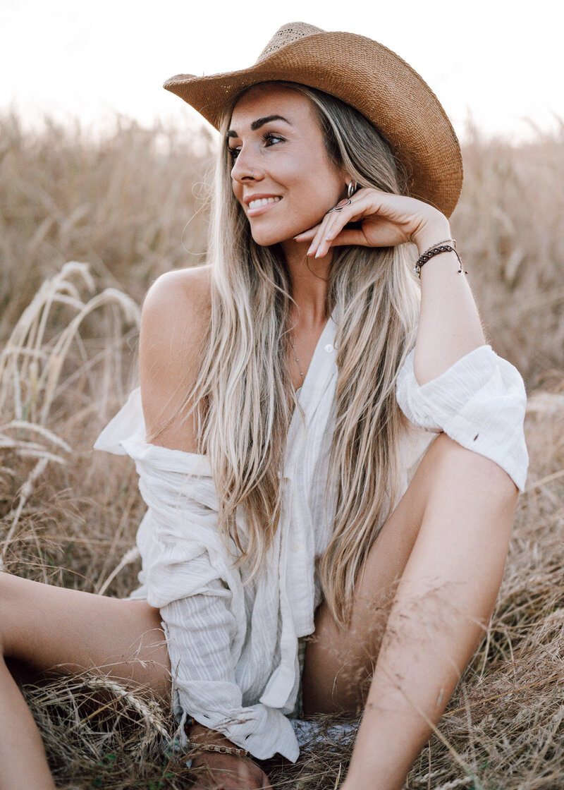 eine blonde Frau sitzt im Feld mit Cowboyhut und hellem Hemd